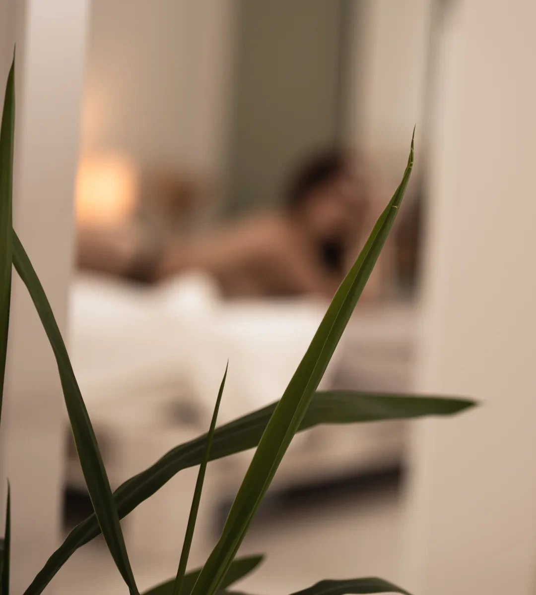 Zurückhaltender Boudoir-Moment hinter einer Pflanze – bewusst nur als leise Andeutung